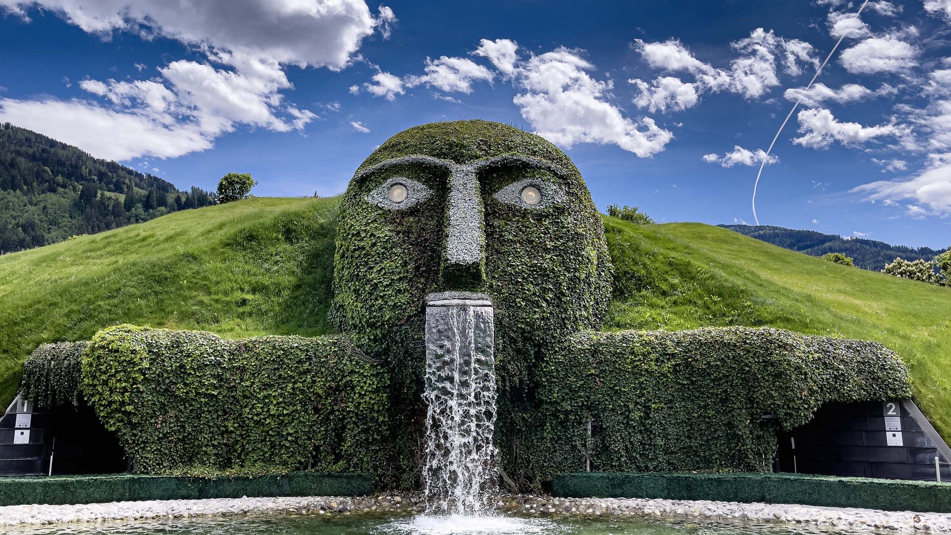 Things to do in Stubaital Green sculpture shaped like a head with water flowing in front of cloudy sky