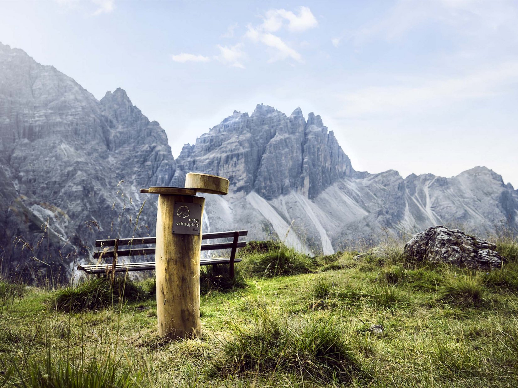 Stubaital: our hotel with a pool Wooden info post with bench in front of rugged mountains and grassy meadow