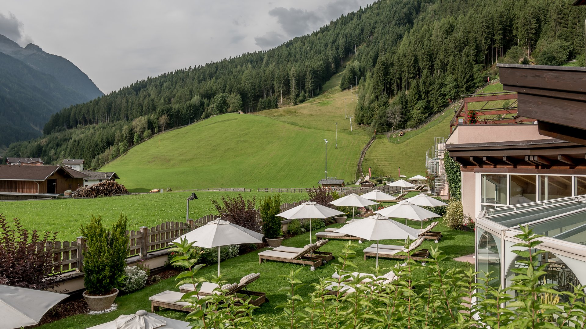 Stubaital: our hotel with a pool Lounge chairs with umbrellas in garden below forested mountain and green hills