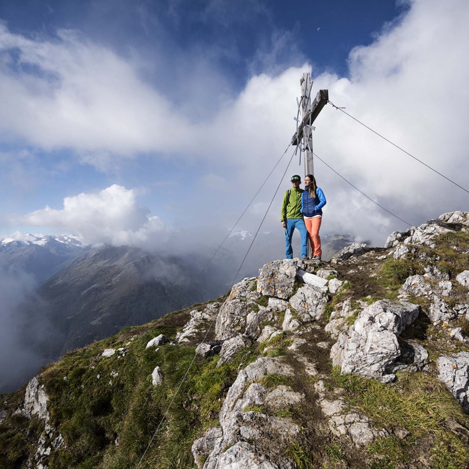 Stubaital: Hiking in summer Two hikers on mountain summit beside large wooden cross under cloudy sky