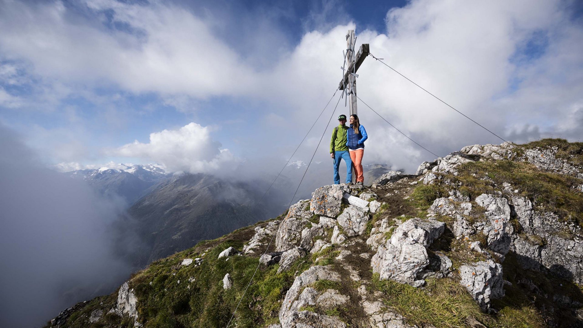 Stubaital: Hiking in summer Two hikers on mountain summit beside large wooden cross under cloudy sky