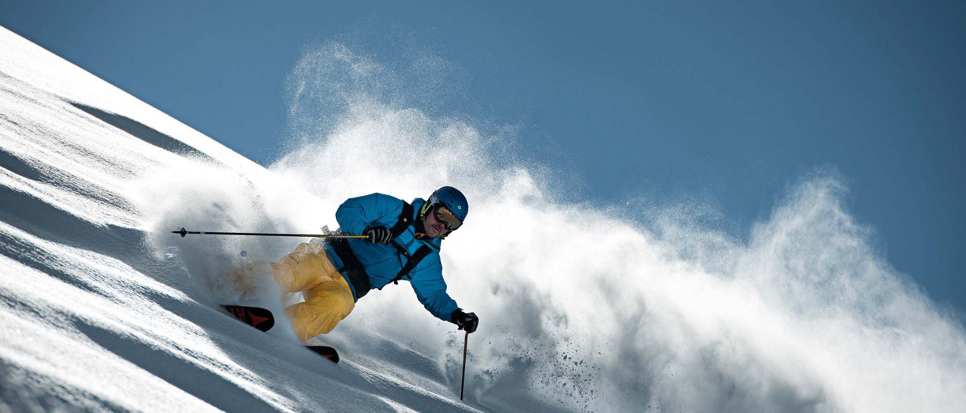 Pisten- und Faschingsgaudi Skifahrer in blauer Jacke und gelber Hose fährt durch Pulverschnee am sonnigen Hang