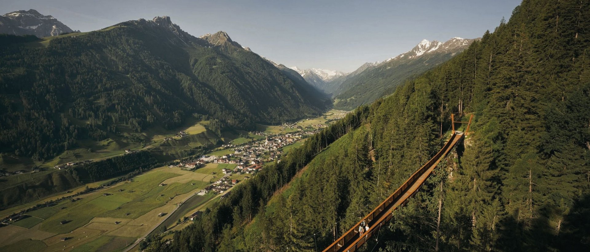 5 Sehenswürdigkeiten im Stubaital, die Sie ganz entspannt erreichen können Luftaufnahme eines Hängebrücke-Wanderwegs im Wald mit Bergtal und Dorf im Hintergrund