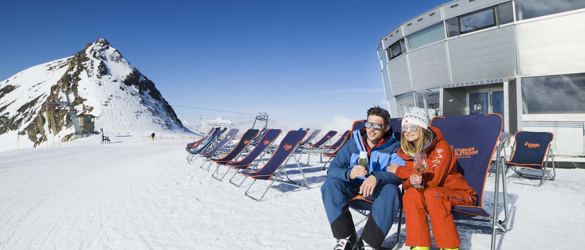 Unser Winter-Special für Sie Paar in Skibekleidung sitzt mit Getränken auf Liegestühlen im Schnee vor Berg