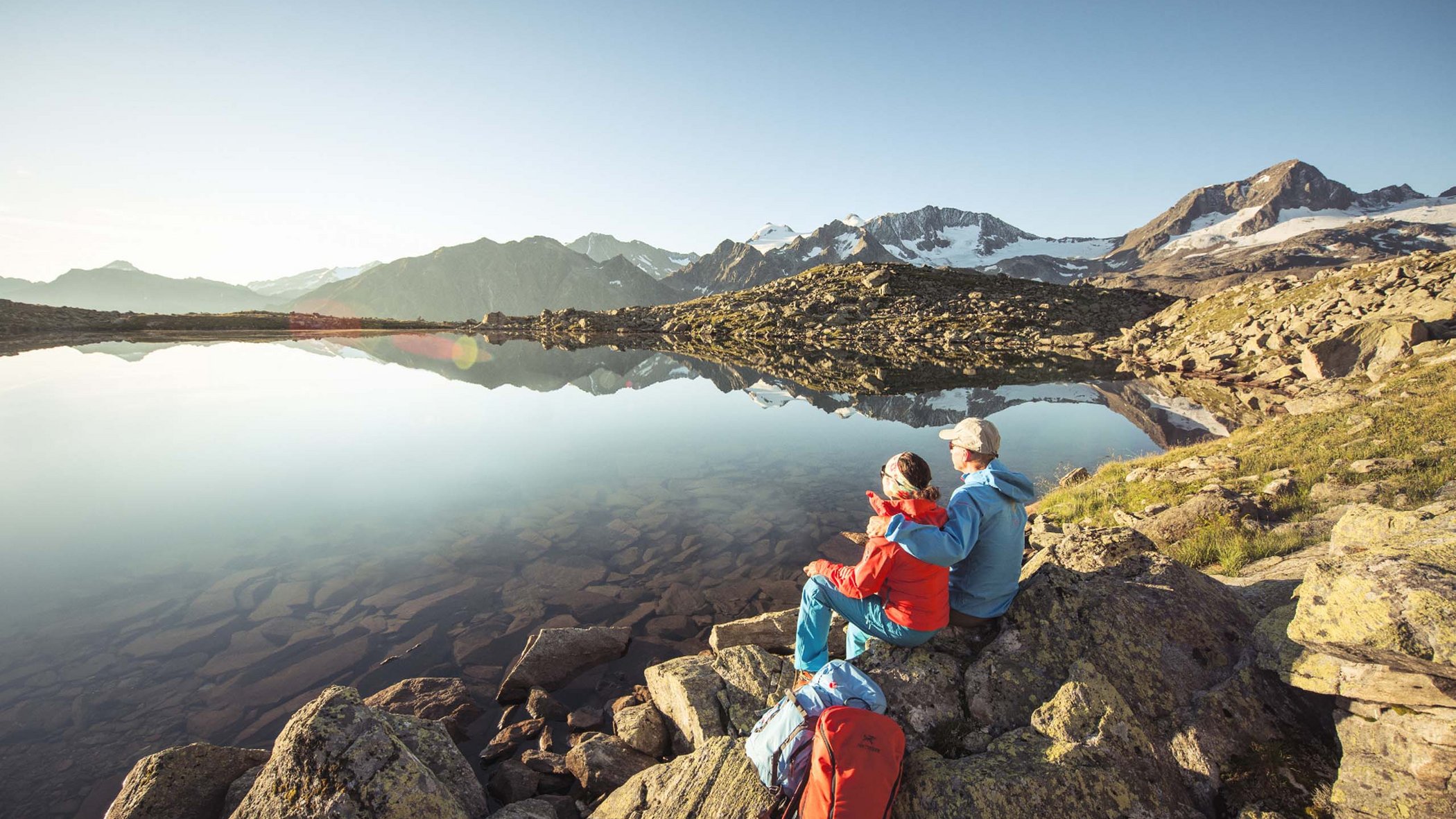 Stubaital: Hiking in summer Two hikers sit on rocks overlooking a calm mountain lake at sunrise